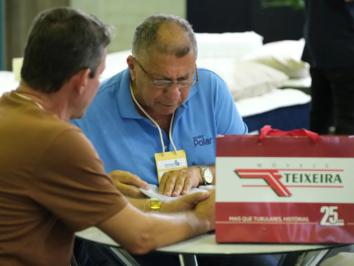 Comprador e representante negociam à mesa, com a sacola da Teixeira ao lado