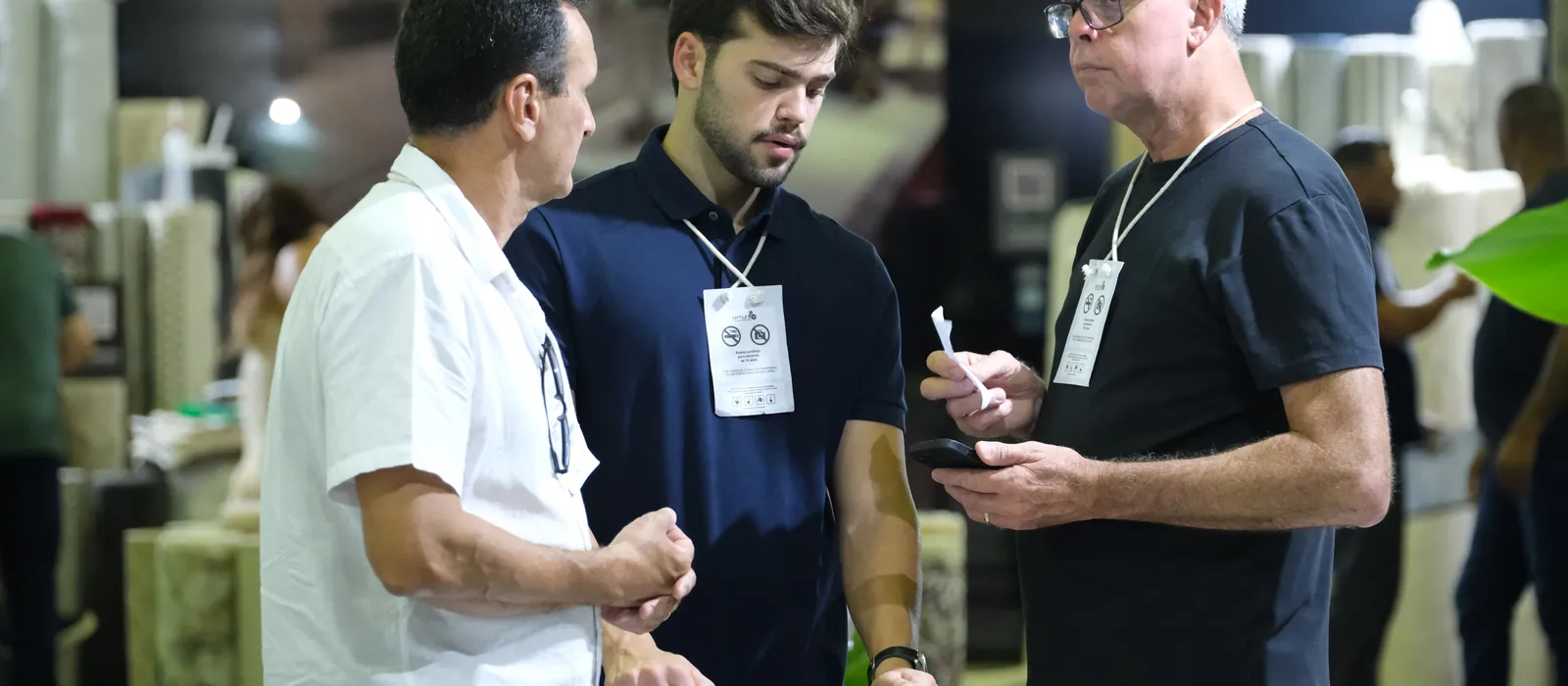 Três homens conversam num estande da FEMUR 2026, um deles com o celular na mão