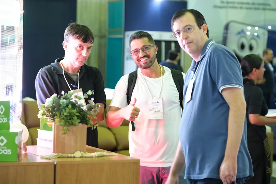 Três visitantes conversam num estande, um deles com o polegar para cima