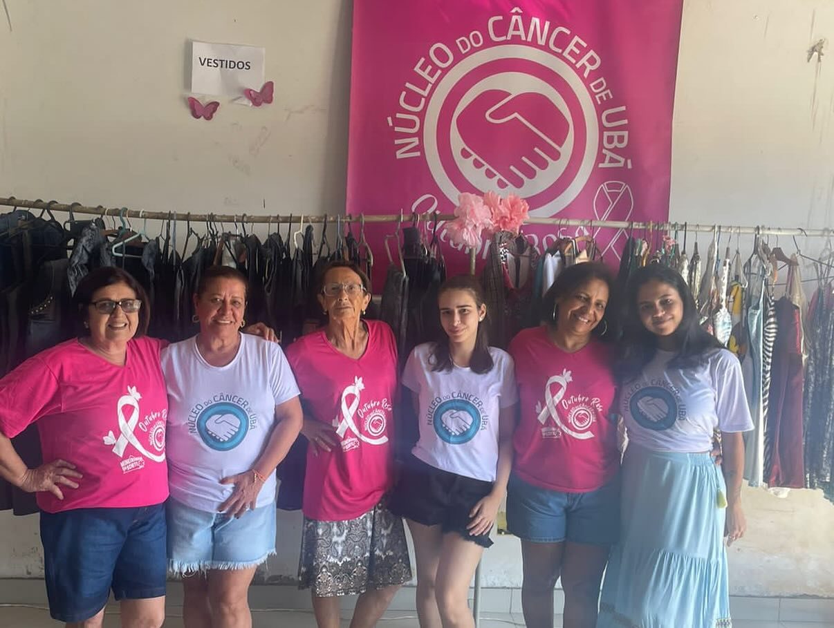 Voluntárias do Núcleo de camiseta rosa diante do painel com o logotipo